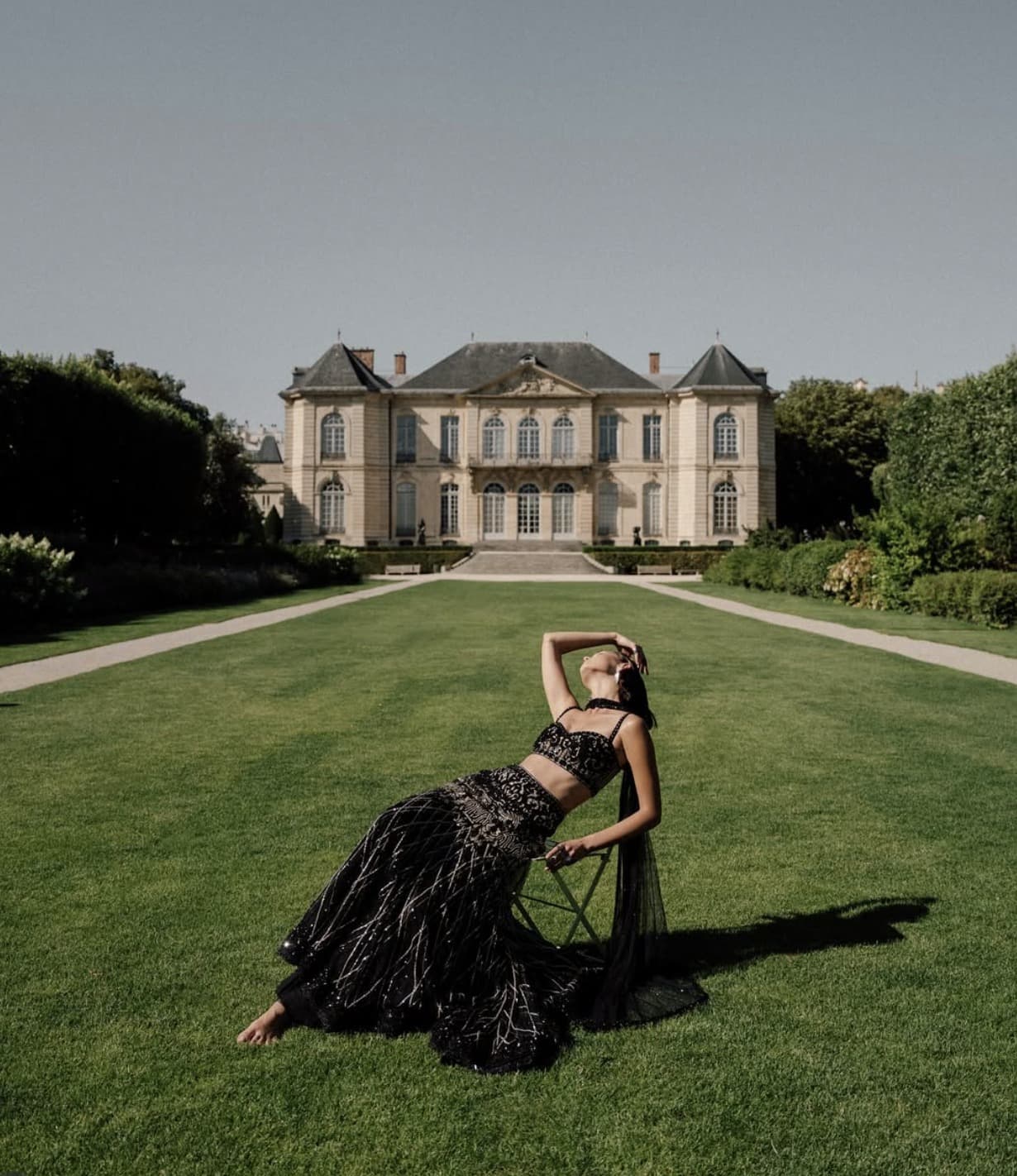 A woman in black reclines on a lawn before a stone house