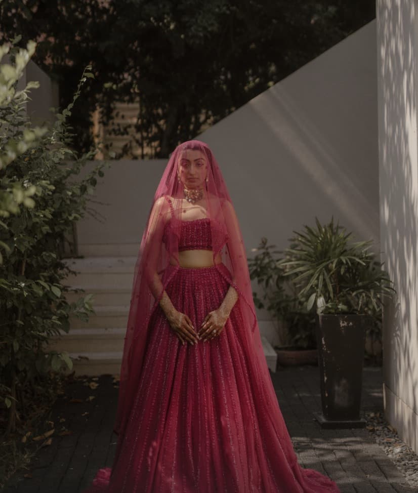 A bride in crimson stands veiled in a shaded garden