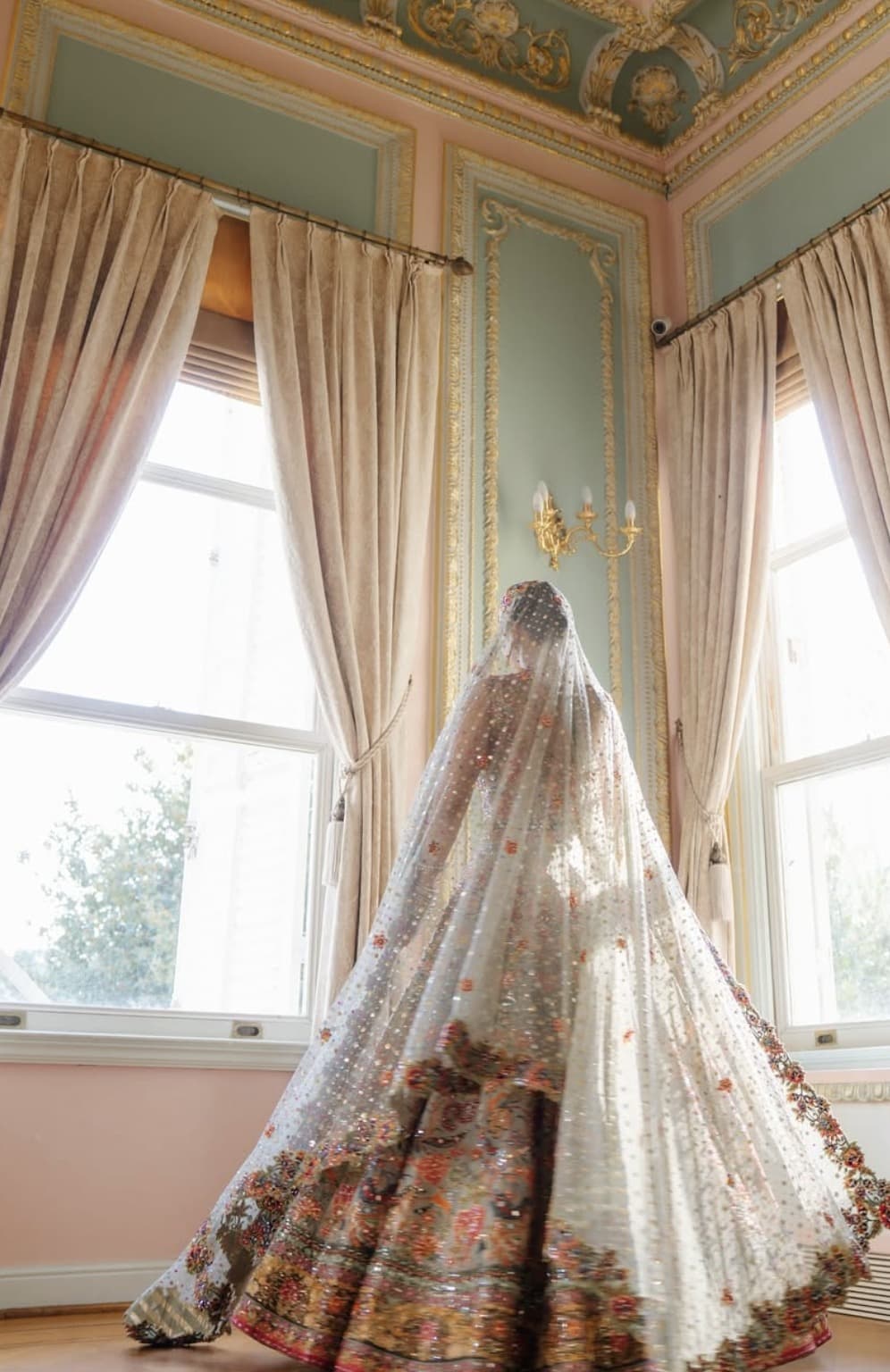 A bride turns beneath a sheer dupatta in a gilded room, sunlight through the windows