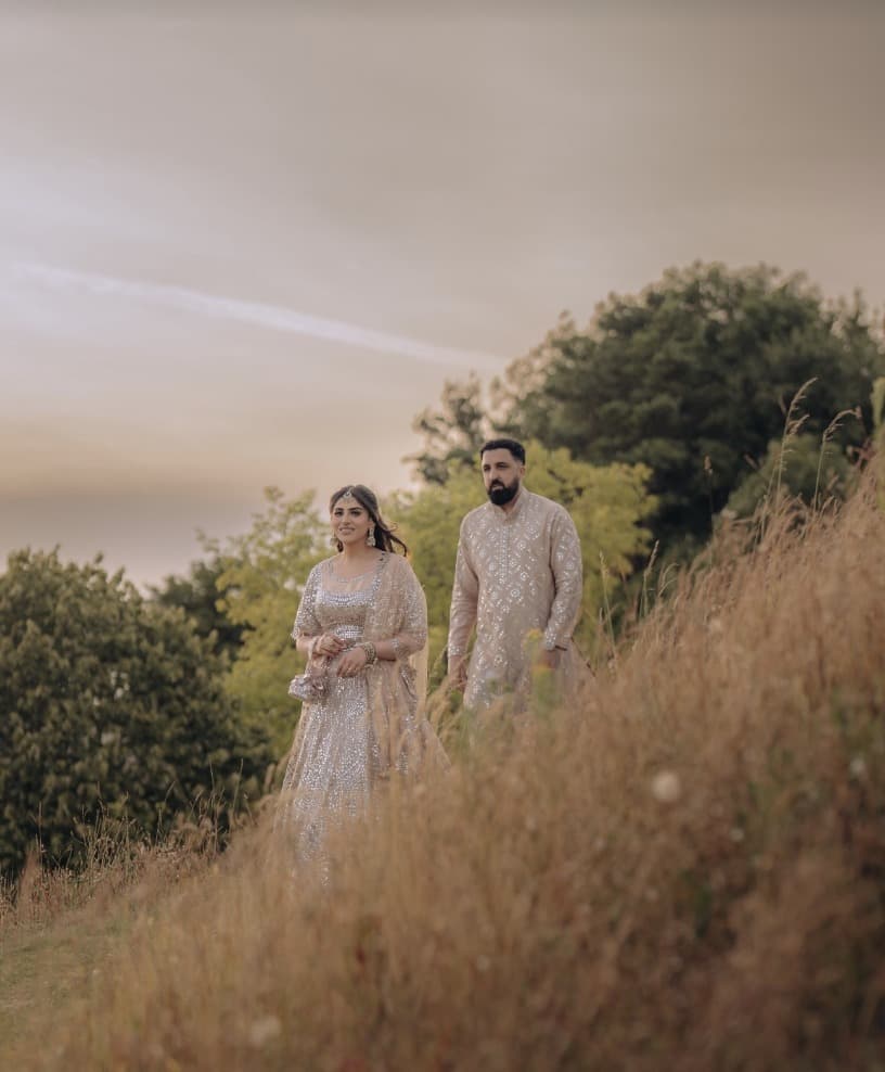 A couple in embroidered ivory walks through tall grass at dusk